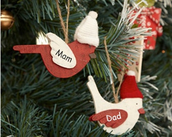 May include: Two bird-shaped Christmas ornaments hanging on a tree. One red bird ornament has the word "Mam" on its wing and a white knit hat. The other is white with "Dad" on its wing and a red knit hat.