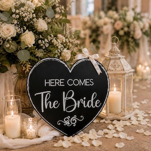May include: A heart-shaped black sign with white text that reads "HERE COMES The Bride" and a decorative swirl at the bottom. The sign is accented with a small white ribbon bow at the top and features a distressed white border around the edges.