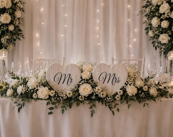 May include: Two white heart-shaped wooden signs with the black script text Mr and Mrs, nestled within a lush floral arrangement of white roses, baby's breath, and green eucalyptus leaves, displayed on a table with white linens and candles.