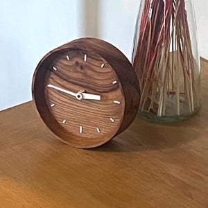 Peut inclure: Une horloge ronde en bois avec un cadran clair et des aiguilles blanches est posée sur une surface en bois. Derrière, un vase en verre transparent rempli de fleurs séchées rouges. C'est un élément de décoration.