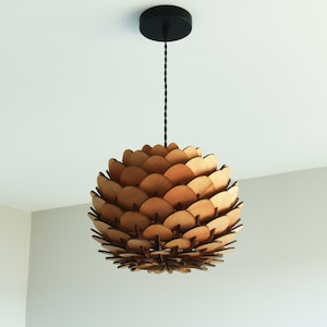 Peut inclure: Un luminaire suspendu en bois avec un design rond et géométrique ressemblant à une pomme de pin. Le luminaire est composé de plusieurs couches de bois, créant un look unique et moderne.