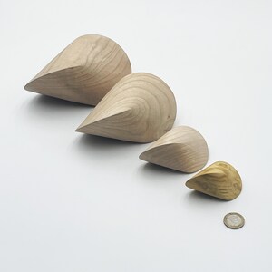 Può includere: Quattro oggetti in legno a forma di goccia di diverse dimensioni, disposti diagonalmente su una superficie bianca. I tre più grandi sono di colore chiaro, mentre il più piccolo ha una tonalità dorata più scura. Una moneta è accanto all'oggetto più piccolo.