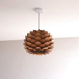 May include: A wooden pendant light fitting with a unique design resembling a pine cone. The light fitting is made of multiple layers of wood, creating a layered effect. The light fitting is suspended from a black cord and a white ceiling mount.