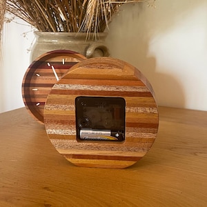 May include: A round wooden clock with a layered wood design in shades of brown, red, and yellow. The clock face is visible through a square opening in the wood. A battery is visible in the clock's compartment.