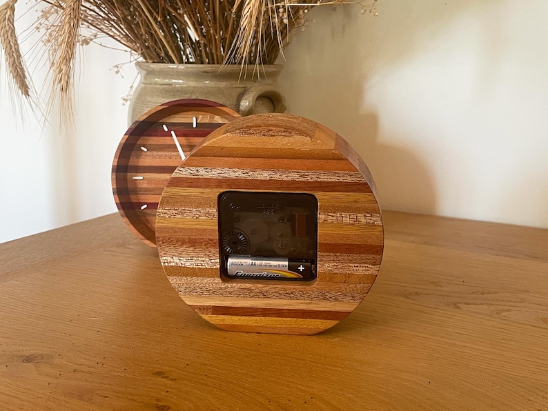 May include: A round wooden clock with a layered wood design in shades of brown, red, and yellow. The clock face is visible through a square opening in the wood. A battery is visible in the clock's compartment.