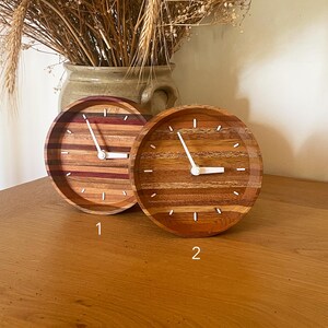 May include: Two round wooden clocks with white hands and Roman numeral markings. The clocks are made of different types of wood, creating a unique and natural look. The clock on the left has a more colorful wood pattern, while the clock on the right has a more uniform wood pattern.