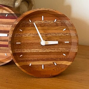 May include: A round wooden clock with white hands and white markings on a wood grain background.