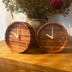 May include: Two wooden clocks with white hands and a striped wood design. The clocks are on a wooden surface.