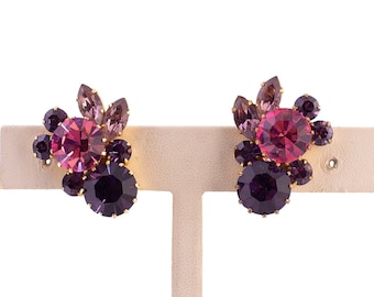 Pode incluir: Um par de brincos vintage de encaixe com um conjunto de strass roxos e rosa. Os brincos têm uma moldura dourada e são exibidos em um suporte creme sobre um fundo branco. O design inclui pedras de várias formas e tamanhos.