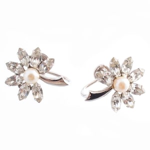 May include: A pair of silver tone clip-on earrings with a flower design. Each earring features a cluster of clear rhinestones surrounding a single faux pearl.