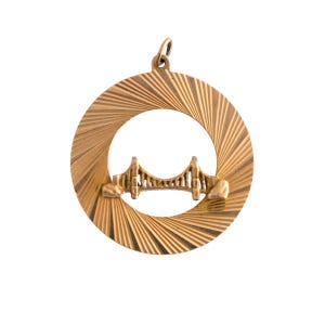 May include: A gold-colored pendant featuring a bridge design within a circular frame. The bridge is detailed with support structures and a connecting roadway. The frame has a radial pattern, and the pendant includes a loop for a chain.