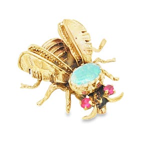 May include: A gold-toned bee brooch with intricate detailing. The bee features an oval, light blue gemstone and two small, round red gemstones. The wings and body have textured patterns, and the legs are delicate.
