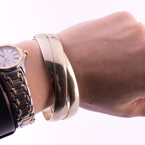 May include: A set of three silver bangle bracelets worn on a wrist. The bracelets are stacked on top of each other and are made of a smooth, shiny metal.