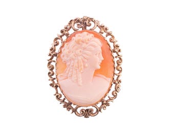 May include: Oval cameo brooch featuring a profile of a woman carved in light pink and orange hues. The cameo is set in a gold-toned filigree frame with intricate scrollwork, creating an elegant and vintage aesthetic.