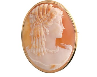 May include: Oval cameo brooch featuring a profile of a woman with detailed hair and a pearl necklace. The cameo is set in a gold-colored frame. The background is a peach color.