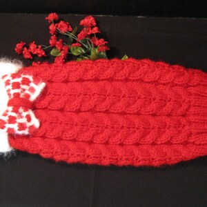 May include: Red cable-knit dog sweater with a white and red checkered bow. The sweater has a white fluffy collar. A sprig of red flowers is placed behind the sweater. The sweater is designed for a small dog.