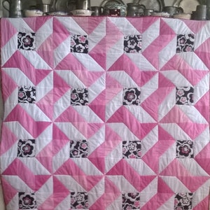 May include: A pink and white quilt with a geometric pattern of squares and triangles. The quilt has a black and white floral print in the centre of each square.