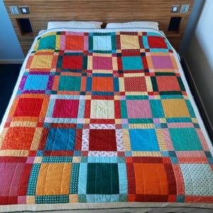 May include: A colorful patchwork quilt with a grid pattern of squares in various colors, including red, orange, yellow, pink, and blue. The quilt is on a bed with a wooden headboard. The quilt's edges are bordered with a cream-colored fabric.