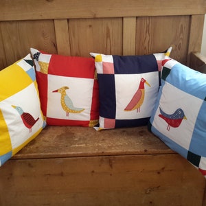 May include: Four colorful patchwork pillow covers with embroidered birds. The pillows are arranged on a wooden bench. The pillows are yellow, red, blue, and navy blue.