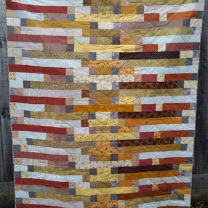 Puede incluir: Una colcha de retazos con un patrón de rayas horizontales en tonos marrones, amarillos y rojos. La colcha está hecha de varias telas con diferentes patrones y texturas.