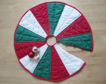 May include: A red, white, and green quilted Christmas tree skirt with a scalloped edge. The skirt is decorated with white polka dots and has a hole in the center for the tree trunk.