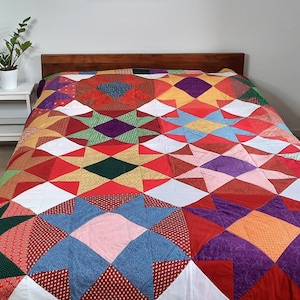May include: A colorful patchwork quilt with a variety of geometric patterns in shades of red, green, purple, blue, and yellow. The quilt is made up of squares and triangles, and it is laid out on a bed.
