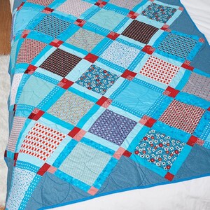 May include: A patchwork quilt with a variety of patterned squares in shades of blue, red, and white. The quilt features a light blue background with red corner accents and a darker blue border. The quilt is made of fabric.