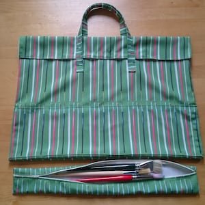 May include: Green striped canvas tote bag with a zippered pocket on the bottom. The pocket is open and contains several paint brushes.