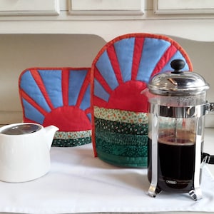 May include: Two quilted pot holders with a sunrise design in shades of blue, red, and green. A silver french press coffee maker with a black handle sits on a white surface.