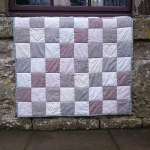 May include: A patchwork quilt with a checkered pattern in shades of gray, white, and pink. The quilt has a teal border and features small heart designs throughout.