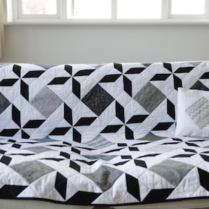 Peut inclure: Une couverture et un coussin géométriques noirs, blancs et gris recouvrent un canapé blanc. La couverture présente un motif répétitif de losanges et d'étoiles, créant un design graphique moderne. Le coussin a un motif en losange.