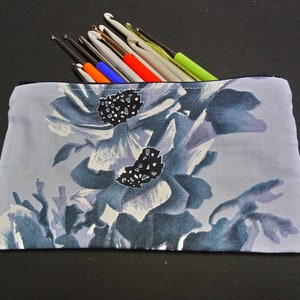 May include: A gray zippered pouch with a floral print featuring blue and white flowers with black centers. The pouch is filled with colorful crochet hooks.