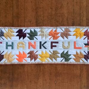 May include: A white quilted table runner with a brown, orange, and yellow leaf pattern. The word "THANKFUL" is spelled out in the centre of the runner using the leaf pattern.