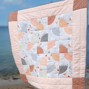 May include: A pink and gray baby quilt with a pattern of curved lines and small shapes. The quilt is made of soft fabric and is perfect for cuddling.