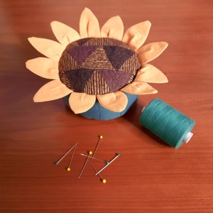 May include: A sunflower-shaped pincushion with a quilted fabric center and orange petals. A spool of teal thread and several sewing pins are on a wooden surface.