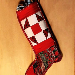 Peut inclure: Une chaussette de Noël rouge et blanche en patchwork avec des étoiles dorées autour du bord. La chaussette est remplie de cadeaux emballés et est accrochée à une surface en bois.