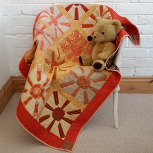 May include: A patchwork quilt with a red border and a variety of orange and yellow patterns. A teddy bear is sitting on the quilt.