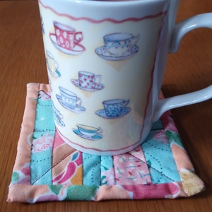 Pode incluir: Uma caneca de cerâmica branca com alça, com um padrão de xícaras de chá e pires em várias cores. A caneca está sentada em um descanso de copo de tecido patchwork colorido.