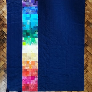 May include: A large, handmade quilt with a vibrant, colorful strip down the center. The strip features a gradient of colors, from purple to red, made of small, patchwork squares. The background is a solid navy blue.