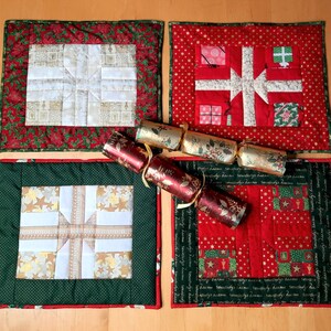 May include: Four red and green quilted placemats with a white, gold, and red patchwork design. The placemats feature a repeating pattern of a wrapped gift box. The placemats are decorated with the text "merry christmas" repeated multiple times.