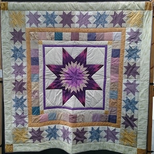 Może przedstawiać: Kołdra w kolorze fioletowym i białym z dużym wzorem gwiazdy w środku. Kołdra jest otoczona obramowaniem z mniejszych gwiazd w odcieniach fioletu, błękitu i żółci.