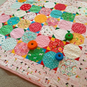 Può includere: Un quilt colorato a patchwork con una varietà di tessuti e motivi. Il quilt è composto da esagoni e quadrati, e presenta una varietà di colori, tra cui rosso, blu, verde, giallo e rosa. Ci sono tre bobine di filo sul quilt.