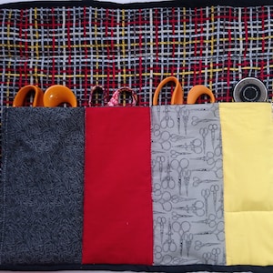 Op de afbeelding: Een zwarte stoffen rolhoes met meerdere vakken om scharen en andere naai-accessoires in op te bergen. De hoes heeft een geruit patroon aan de buitenkant en verschillende stoffen met patronen aan de binnenkant.