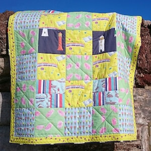 May include: A patchwork quilt featuring a mix of colourful fabrics, including yellow, blue, green, pink and red. The quilt is made up of squares and rectangles, and features a variety of designs, including ice cream cones, lighthouses and boats. The quilt is hanging on a stone wall.
