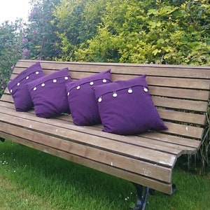 May include: Four square, purple throw pillows with decorative silver-colored buttons are arranged on a wooden bench. The pillows are made of a solid, dark purple fabric. The bench is outdoors, with green grass and foliage in the background.