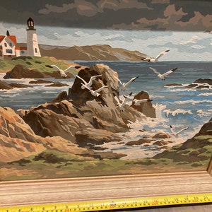 May include: A framed painting of a seascape with a lighthouse in the distance. The painting features a rocky coastline with crashing waves and seagulls flying overhead. The sky is a cloudy blue and the water is a deep blue.