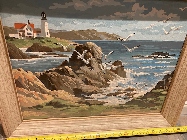 May include: A framed painting of a seascape with a lighthouse in the distance. The painting features a rocky coastline with crashing waves and seagulls flying overhead. The sky is a cloudy blue and the water is a deep blue.