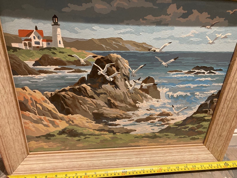 May include: A framed painting of a seascape with a lighthouse in the distance. The painting features a rocky coastline with crashing waves and seagulls flying overhead. The sky is a cloudy blue and the water is a deep blue.
