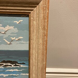 May include: A close-up of the top right corner of a wooden frame. The frame is light brown with a white wash and has a slightly distressed look. The frame is surrounding a painting of a seascape.