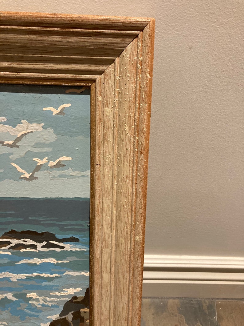 May include: A close-up of the top right corner of a wooden frame. The frame is light brown with a white wash and has a slightly distressed look. The frame is surrounding a painting of a seascape.